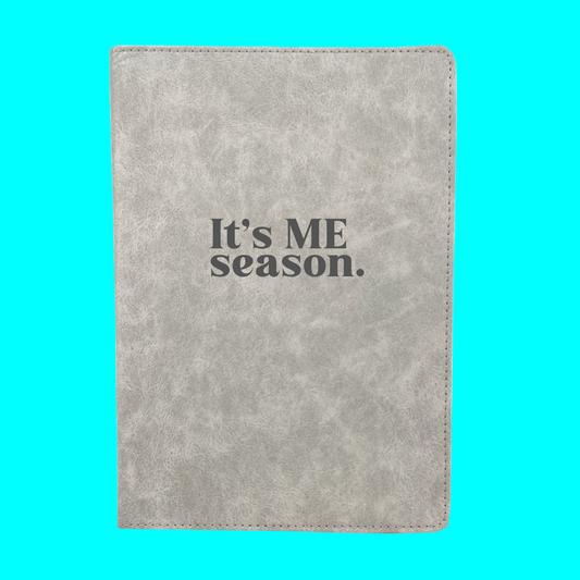 Gray notebook with 'It's ME season.' text on a turquoise background
